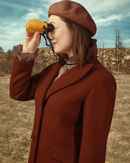 A woman in a beret and coat looks through yellow binoculars in a grassy field with bare trees and a blue sky, vintage-style photography.